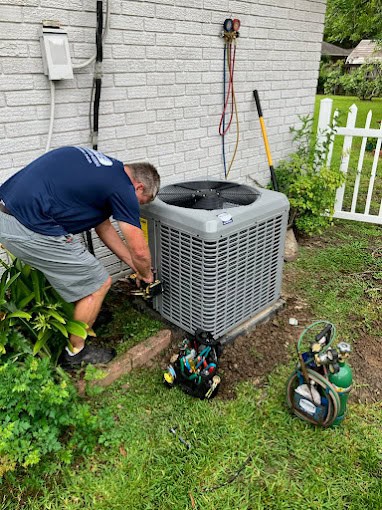 A technician from an HVAC company in Baton Rouge is repairing or installing an air conditioning unit outside a building. Wearing a blue shirt and shorts, he is surrounded by tools and equipment, with a white brick wall and greenery in the background.