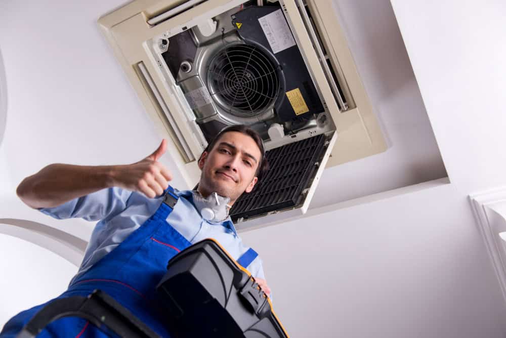 A maintenance worker in blue overalls stands on a ladder, giving a thumbs-up while holding a toolbox. He is positioned under an open ceiling air conditioning unit. The perspective is from below, looking up towards the worker and the unit.